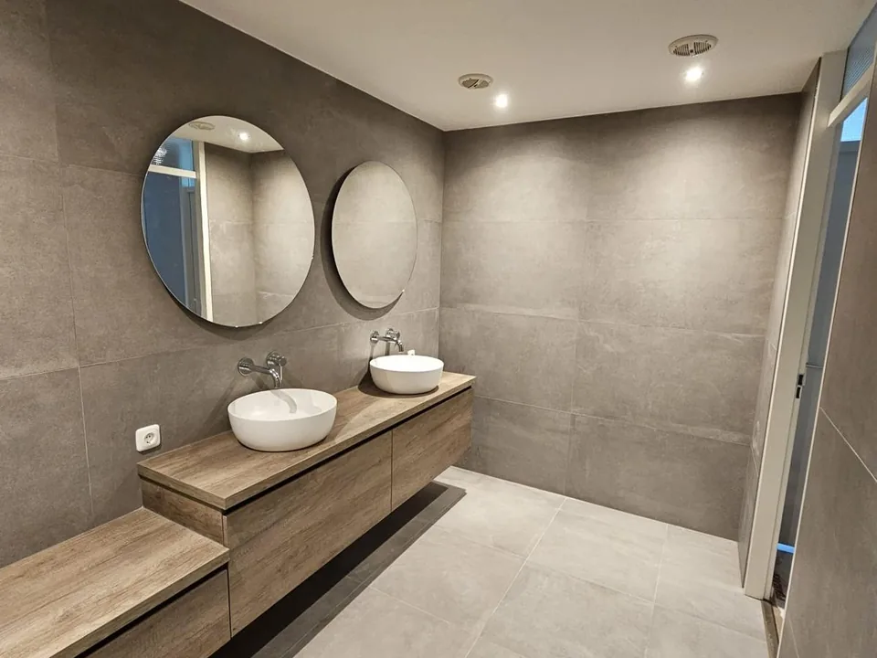 Badkamer met grijze betonlook tegels, zwevend eiken badmeubel en twee ronde spiegels
